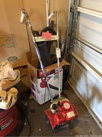 Rototiller unit sitting on floor next to box featuring a tiller image, showing red body and metal handles with control wires attached.