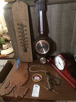 Full lot photo showing wooden owl carving, SkyTimer mantle clock, vintage barometer with attached thermometer, vertical wooden thermometer, two bolo ties, and auction tag.