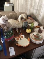 Photo of a collection of bird and animal figurines on a table including an owl made of shells, colored glass owl, ceramic owl dish, wooden and ceramic duck and goose figures, a green glass item, a small yellow pumpkin decor, decorative plates, and various small owl ornaments.