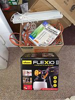 Boxed Wagner Flexio 2000 paint sprayer, orange extension cord, white task light fixture, and halogen bulb packages in a cardboard box.