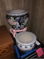 Two decorative ceramic planters shown together; dark floral planter on pink stand and white with blue design planter on blue pad.