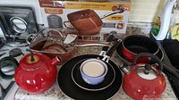 Photo showing red tea kettles, a Copper Chef copper-colored all-around cooking pan, frying pans, and glass baking dish on kitchen counter.