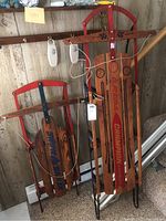 Front view of two wooden snow sleds with metal frames; left sled is blue (Snow Sailer), right sled is red (Reading Champion).