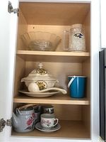 Kitchen cabinet interior shelf containing all items: top shelf holds glass serving bowl and Baltimore Ravens glass mug with ice cubes; middle shelf has ceramic soup tureen with lid and ladle with yellow rose design and a blue container; bottom shelf contains teapot with pink rose floral design and matching teacup with saucer.