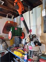 Leaf blower hanging from the ceiling, green Flymo broadcast seeder with red wheels, assorted tools and containers on a workbench