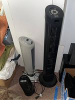 Three tower fans grouped together in a corner: a large black Sunter, medium gray BionAire, and a black LASKO fan.