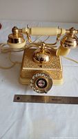 Front view showing ornate gold and cream rotary telephone with pictorial rotary dial and ruler for scale