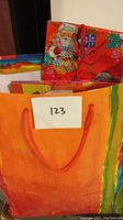 Photo of a large orange gift bag with red rope handles and colorful edge detail, featuring a Santa Claus themed bag behind it.
