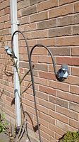 Two metal hanging basket holders with bird decorations seen from a distance mounted on a brick wall near a drainpipe.