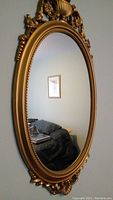 Full view of oval mirror hanging on wall showing detail of gold frame and reflective glass surface.