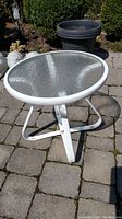 Outdoor round table with textured glass top on white metal tripod base placed on stone patio.