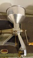 Close-up of the hand cranked tomato machine showing metal funnel, crank handle, and mesh filter.