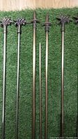 Six metal drapery rods laid on grass, showing decorative flower and fleur de lis finials, pewter and bronze finishes, various lengths.