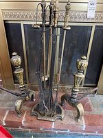 Photo showing the brass andiron pair in front of a fireplace alongside the five-piece fireplace tool set standing in a holder.