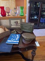 Overview of the lot showing two military hats, two books, a vintage pilot photograph in a frame, and the green ammunition box on a wooden table.