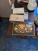 Three music boxes on table showing decorative lids including black lid with gold flowers, cream lid with birds and flowers, wooden box with velvet interior