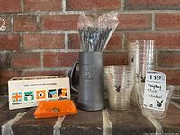 Photo showing tall plastic server jug with embossed Playboy bunny logo, box of Playboy Club matches, orange glass trays with vintage pin-up, clear plastic cups stacked, and plastic stir sticks in jug.