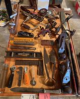 Overview image showing a variety of vintage hand tools laid out on a wooden table including hand planes, hand drills, sharpening stone, levels, and measuring tools.