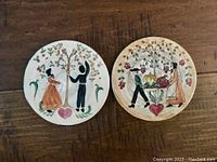 Both pottery plates showing folk art scenes of Amish people with apple tree and harvest, displayed side by side on wooden surface.