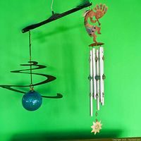 Photo showing two wind chimes against a green background, one black metal spiral with blue glass ball, other Kokopelli with silver chimes and sun pendant