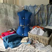 Lot includes blue adjustable dress form, assorted fabrics including plaid shirts, basket with folded fabric and shirts, and white hanging organizer