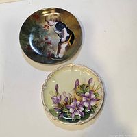 Both plates displayed on a white surface, showing the Grandma's Garden plate with child image and the Bavarian floral plate with scalloped edges together.