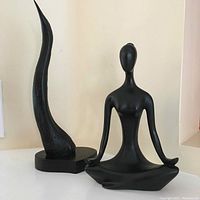 Front view of both abstract black sculptures shown on a white tabletop, one is female figure meditating, other is curved abstract shape on base.
