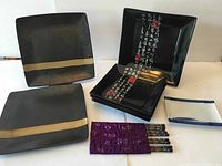 Photo showing black square plates with a gold stripe, glossy black plates with Japanese script, white rectangular plates, and a set of decorated chopsticks with purple cloth holder.