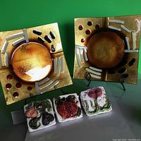 Three square ceramic wall plaques with raised, painted fruit and foliage designs. Two square gold-toned glass art plaques with abstract circular designs with black, white, and red accents. All items arranged on a green background, with glass plaques on stands.