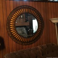Full view of the large round mirror on a wooden paneled wall with brown tufted couch and lamp next to it.