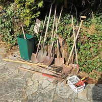 Full view of garden tools lined up on stone patio including shovels, rakes, and hand tool basket.
