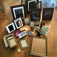 Photo showing 19 assorted photo frames of varying sizes, colors, and designs arranged on wooden floor and table. Includes wood, metal, and decorative styles.