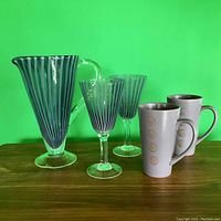 Murano glass pitcher with matching striped glasses and two patterned ceramic mugs on wooden surface against green background