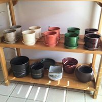 Front view of two shelves showing overall set of 14 pottery planters in various colors and styles on wooden rack. Rack not included.