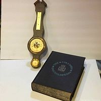 Full view of vintage weather instrument and encyclopedia book together on white surface.
