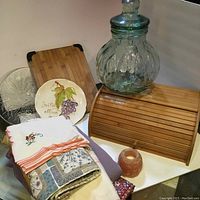 Photo showing wooden bread box, clear textured glass pitcher with lid, two decorative textured glass bowls, hand-painted grape ceramic plate, various folded linens including embroidered and patterned fabrics, and small amber candle holder arranged together.