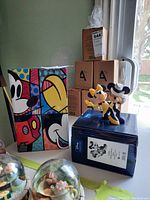 Display of two Disney figurines: a colorful Britto style Mickey Mouse figurine and a black-and-white style Mickey and Minnie Mouse figurine set, each with their original boxes.