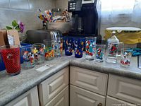 Set of eleven Disney collectible drinking glasses aligned on countertop, featuring various colorful character designs including Mickey Mouse and Donald Duck.