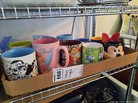 Photo of 10 Disney character mugs lined up in a cardboard box on metal shelf.