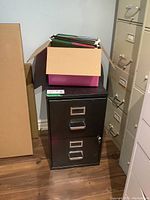 Black two-drawer metal filing cabinet with lock and keys. Cabinet placed on wood floor next to other filing furniture. Box with hanging file folders on top.