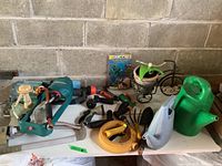 Photo showing assorted garden supplies against cinder block wall including watering cans, water sprinklers, nozzles, and a bicycle-shaped planter
