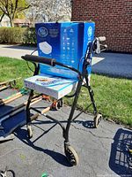 Photo of black metal Health Walker B walker with four wheels and padded handles. On walker seat is a box for Home Care Toilet Safety Seat and a flat box, placed on pavement outdoors with grass and brick building background.