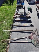 Photo showing nine garden tools including rakes, shovels, pick, hoe, and a green garden cart against a driveway and grass