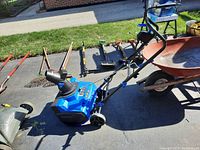 Side view of Snowjoe snow thrower showing blue body, handlebar, and wheels with several garden tools and a wheelbarrow in background.