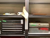 Two metal toolboxes on a shelf. Left is a dark metal toolbox with 4 drawers and handle on top, sticker showing dimensions 22x13x14 and lot tag. Right is a different metal toolbox with latch and stickers, no drawers.