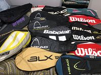 Photo showing multiple tennis racket bags from Wilson, Head, Babolat, and Extender with color variety and brand logos visible.