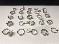 Photo showing 25 assorted stainless steel rings with size tags attached, arranged in rows on white surface.