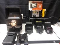 Full view of all vintage camera equipment including cameras, flashes, slide projector, and binoculars arranged on black surface