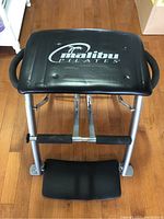 Front view of Malibu Pilates Chair showing padded seat with Malibu Pilates logo and silver metal frame with black foam handles and footrest.