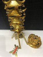 Side view of brass Three Legged Toad pottery with decorative colored stones and brass Feng Shui wind chime with multiple tiers of small hanging bells and ivory-colored hanging elements, sitting on white surface with black background.